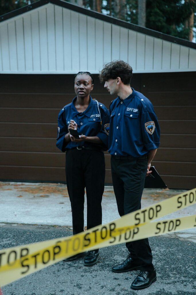 Two police officers standing behind barrier tape in front of a garage, investigating a crime scene outdoors.